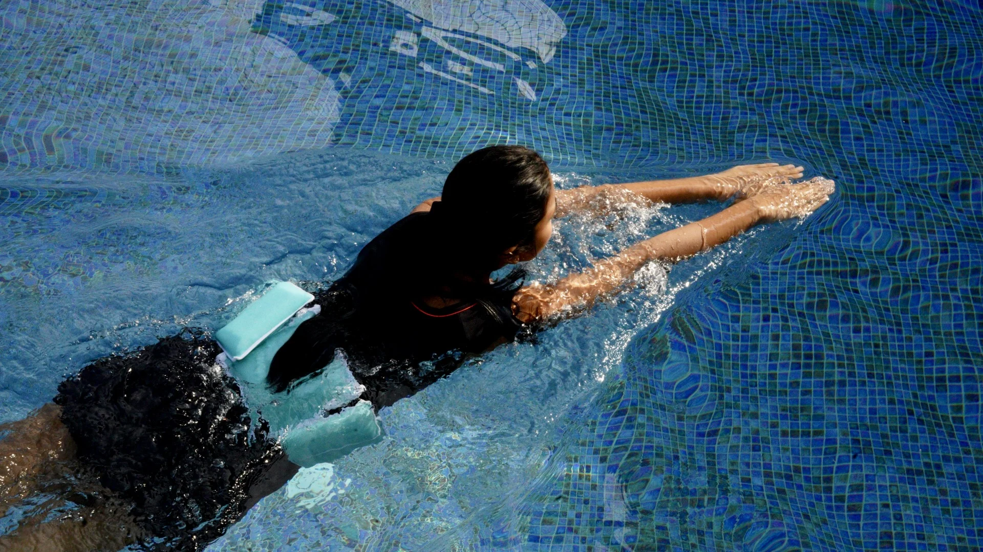 A swimmer doing front crawl in a blue tiled pool, wearing the belt at the lower back